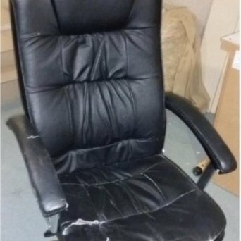 Старое черное офисное кресло из кожи стоит в помещении офиса. У кресла вытерто сидение и требуется замена наполнителя.