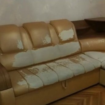 В углу комнаты стоит кожаный угловой светло-коричневый диван. Подголовники и сидения вытерты от частого использования. Подлокотники деревянные.  До ремонта.